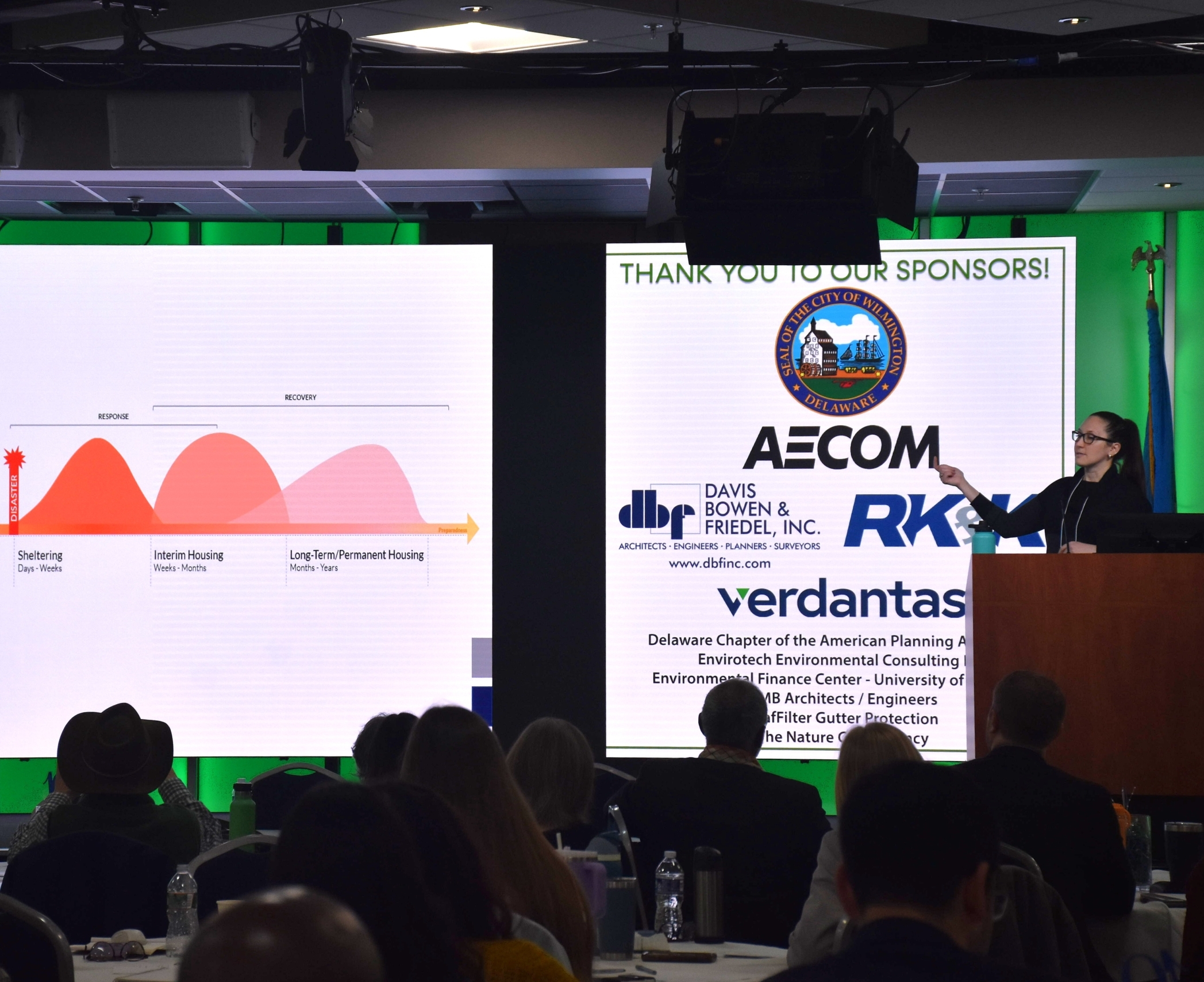A speaker at a podium addressing a seated conference audience, with large screens showing disaster recovery phases and event sponsors
