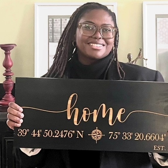 Dominique Bennett smiling and holding a black wooden home sign engraved with the coordinates of her new house