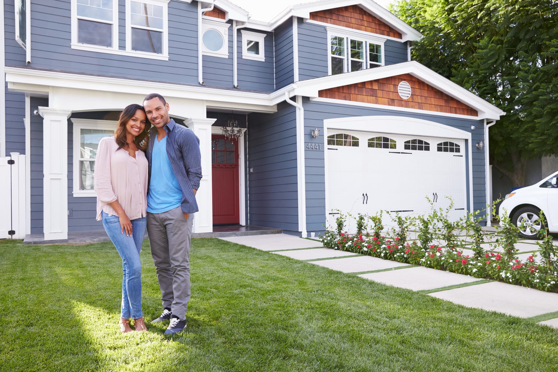 A smiling couple standing on the front lawn of their two-story blue house with a red front door